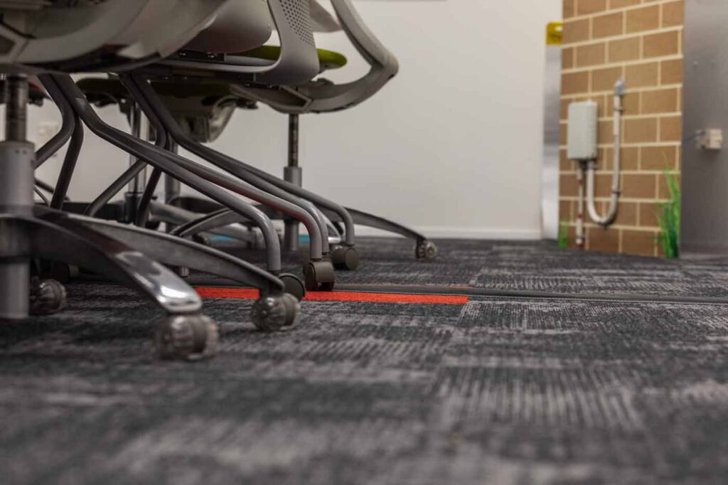 Low-angle view of office chairs on patterned carpet tiles in a busy commercial workspace.